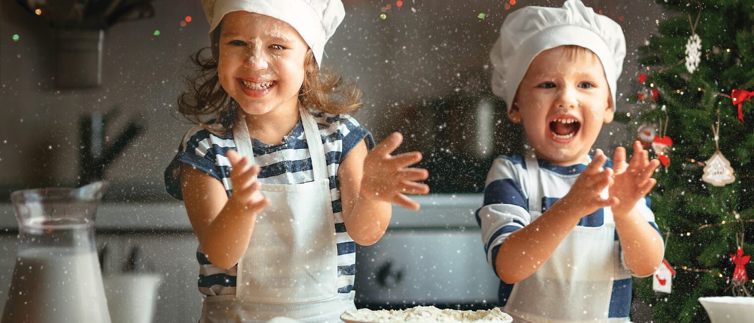 Zwei Kinder backen Weihnachtsgebäck und freuen sich. Im Hintergund ein Weihnachtsbaum in einer Backstube.
