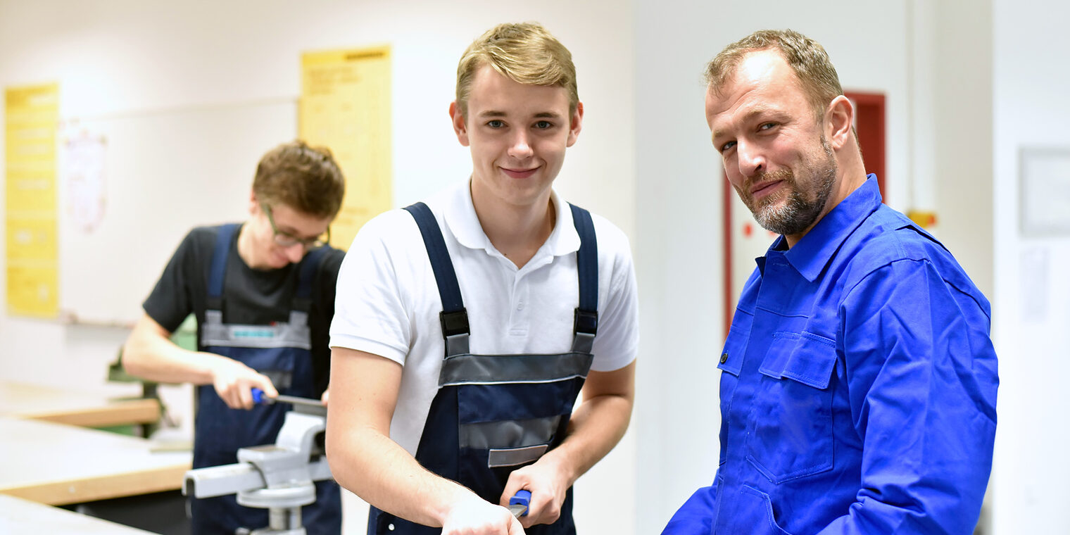berufsausbildung, azubi, ausbildung, handwerk, industrie, mann, personen, jugendlicher, berufsbildung, auszubildenter, beruf, arbeiten, gruppe, arbeitsplatz, kaukasier, jung, arbeitskleidung, lächeln, fröhlichkeit, lachen, professionell, ausbilden, meister, schüler, lehrling, schule, training, lehren, jugendliche, schraubstock, praxis, praktisch, nachwuchs, karriere, berufsschule, bildung, ausbilder, lehrer, azubis, vor ort, qualifizierung, qualifiziert, tätigkeit, metallbau, stahlbau, werkstück, bauteil, berufsbekleidung, werkstatt, werkstätte, Meister Lehrling ÜLU Überbetriebliche Lehrlingsunterweisung